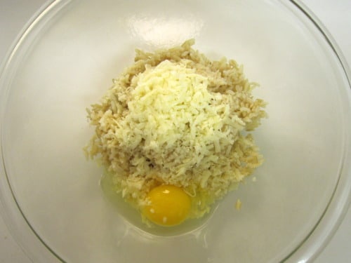 Glass bowl with cooked brown rice, shredded cheese, and an egg, prepared for a brown rice crust.