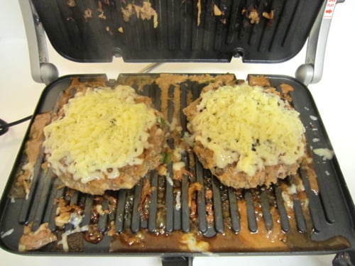 Bulgogi cheeseburgers cooking on an indoor grill, topped with melted cheese and surrounded by savory juices.