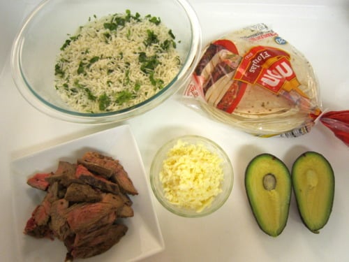 Ingredients for carne asada tacos, including sliced grilled steak, cilantro rice, shredded cheese, flour tortillas, and a halved avocado, arranged on a white surface.