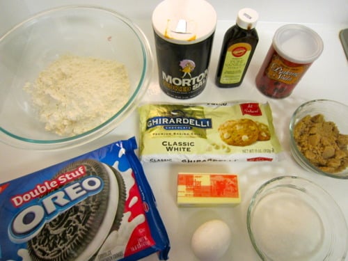 Ingredients laid out on a counter, including flour, salt, vanilla extract, baking soda, white chocolate chips, brown and white sugar, butter, an egg, and a package of Double Stuf Oreos.
