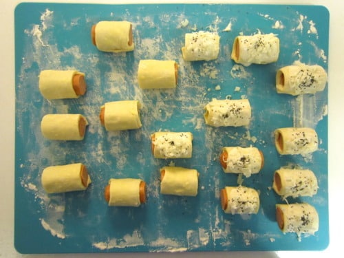 Unbaked pigs in a blanket arranged on a floured blue cutting board, some topped with shredded cheese and herbs.