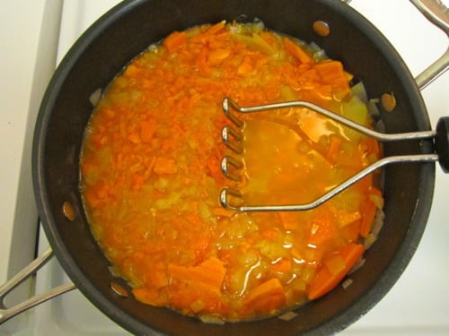 Diced sweet potatoes being mashed in a pot with sautéed onions and broth, prepared for sweet potato and spicy sausage soup.