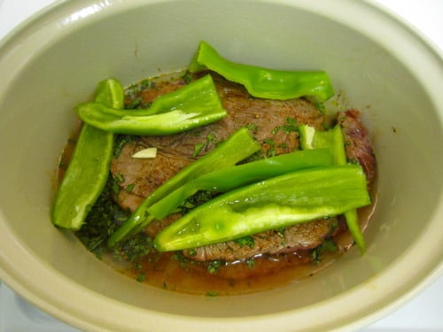 Beef barbacoa cooking in a pot with sliced green peppers, garlic, and herbs.