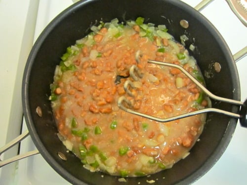 Pinto beans being mashed in a skillet with sautéed onions and green bell peppers, prepared for refried beans in eggs barbacoa.