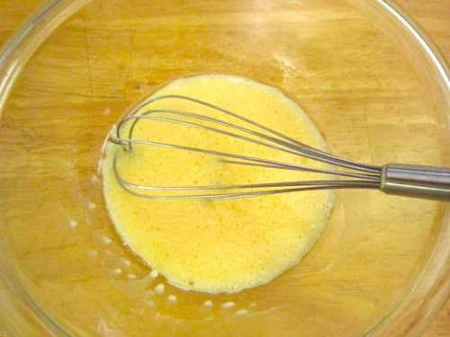 Whisk mixing eggs and batter in a glass bowl for blueberry lemon cream cheese muffins.