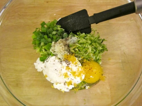 Glass bowl with ingredients for zucchini ricotta fritters, including shredded zucchini, ricotta, egg, chopped green pepper, garlic, lemon zest, and black pepper.