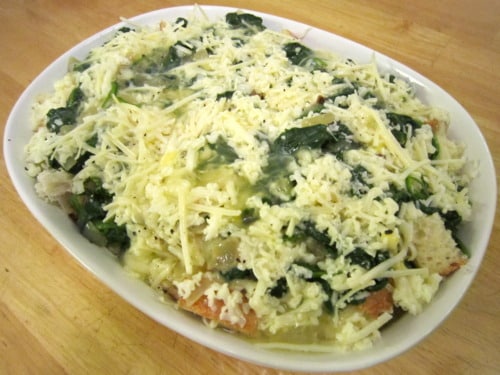 Unbaked spinach and cheese strata in a white baking dish, layered with bread, sautéed spinach, and shredded cheese.