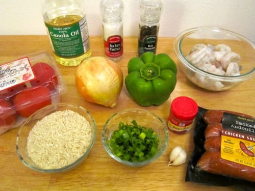 Ingredients for shrimp jambalaya, including tomatoes, rice, onion, green bell pepper, green onions, garlic, shrimp, chicken andouille sausage, canola oil, seasonings, and spices, arranged on a wooden surface.