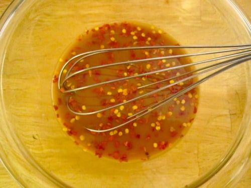 Whisked stir fry sauce in a clear glass bowl with red pepper flakes, shown being mixed with a metal whisk on a wooden surface.