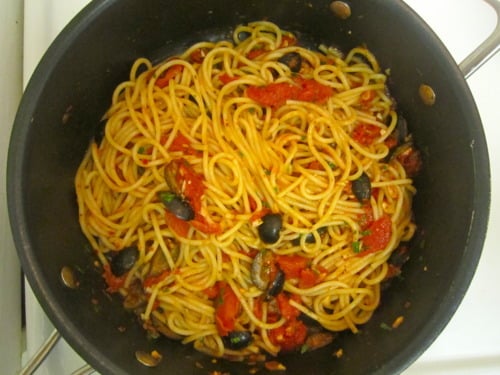 Spaghetti alla puttanesca in a pot with tomatoes, black olives, capers, and herbs.