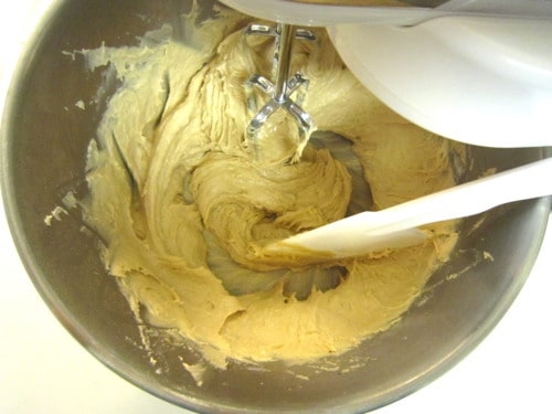 Cookie dough being mixed in a stand mixer bowl with a paddle attachment and white spatula, showing a thick, creamy texture
