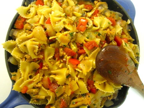 Skillet lasagna made with bowtie pasta, ground meat, diced red peppers, onions, and herbs in a blue skillet with a wooden spoon.