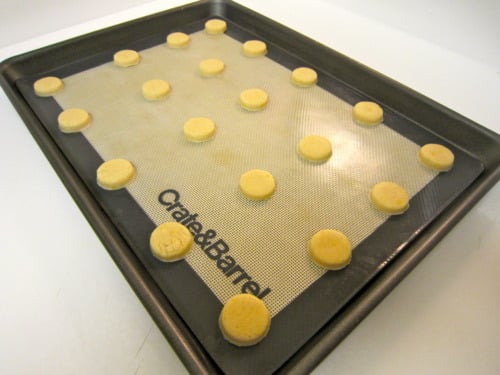 Unbaked lemon cookie rounds on a silicone baking mat-lined baking sheet, prepared for mini strawberry-filled sandwich cookies.