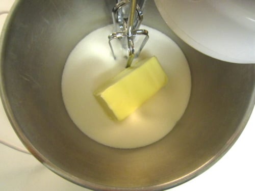 A stick of butter and granulated sugar sit together in a metal mixing bowl beneath an electric mixer’s beaters, ready to be creamed.