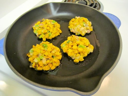 Four corn cakes frying in a skillet with visible corn kernels and diced vegetables.