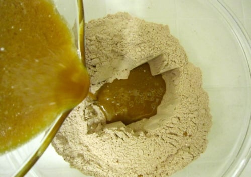Wet mocha mixture being poured into a bowl of dry chocolate muffin mix in a clear mixing bowl.