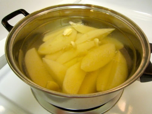 Peeled potato wedges simmering in a pot of water with garlic cloves on a stovetop.