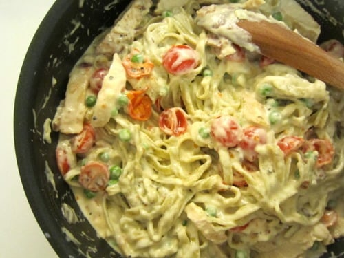 Creamy chicken Alfredo pasta with cherry tomatoes and green peas being stirred in a pan.”