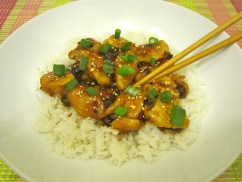 General Tso's chicken served over white rice, garnished with sesame seeds and chopped green onions, with chopsticks.