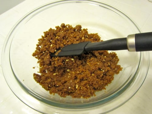 Glass bowl with graham cracker crust mixture being stirred using a black spatula.