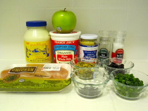Ingredients for chicken salad including cooked chicken breast, mayonnaise, Greek yogurt, apple, dried cherries, pistachios, green onions, and seasonings arranged on a kitchen counter.