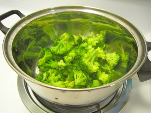 Fresh broccoli florets being steamed in a stainless steel pot on the stovetop for pad see ew.