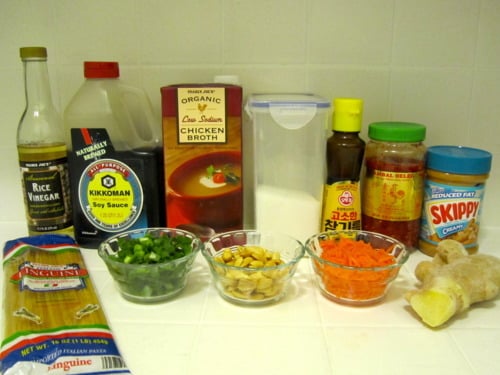 Ingredients for vegetarian peanut pasta, including linguine, rice vinegar, soy sauce, chicken broth, sugar, sesame oil, chili garlic sauce, peanut butter, ginger, green onions, peanuts, and shredded carrots, arranged on a countertop.