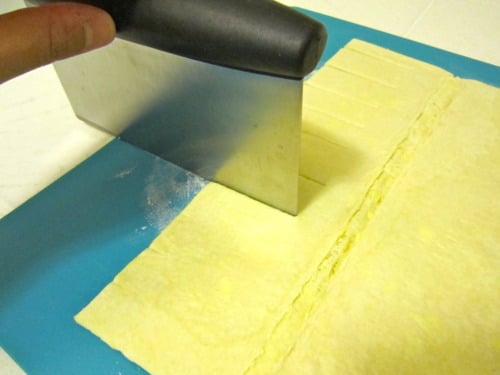 Puff pastry sheet being cut into strips on a blue cutting board using a bench scraper, prepared for orange cheese danish.