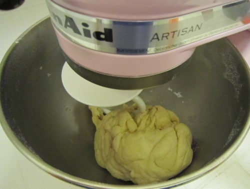 Pizza dough being kneaded in a stand mixer with a dough hook attachment.