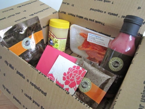 Open cardboard shipping box containing assorted food items and packaged ingredients, including chocolate-covered treats, a bottled sauce, wrapped meat, and a greeting card.