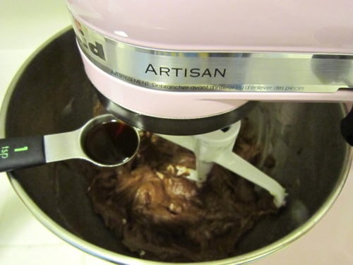 Stand mixer blending chocolate cookie dough while a measuring spoon adds vanilla.