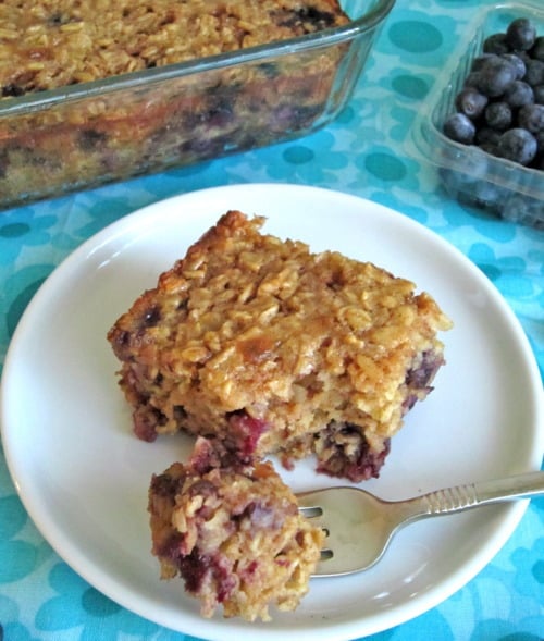 Square slice of baked blueberry oatmeal on a white plate with a fork, with a larger dish of the oatmeal and fresh blueberries in the background.