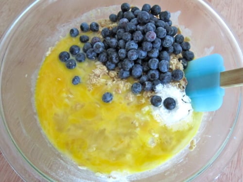 Mixing bowl with oats, fresh blueberries, flour, and melted butter being combined with a blue spatula.