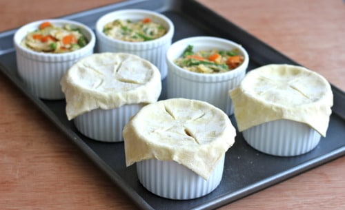 Individual chicken pot pies in white ramekins on a baking sheet, with puff pastry crusts placed on top and ready for baking.