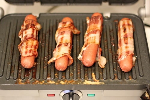 Bacon wrapped hot dogs cooking on an electric grill.