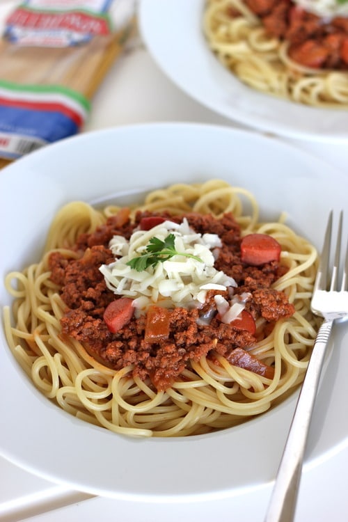 Pinoy-style spaghetti with ground meat, sliced hot dogs, and tomato sauce, topped with shredded cheese and parsley, served on a white plate with a fork.