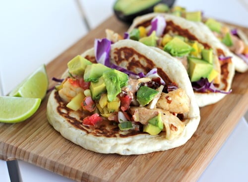 Biscuit-style lime fish tacos filled with flaky fish, diced avocado, fresh salsa, and purple cabbage, served on a wooden board with lime wedges.