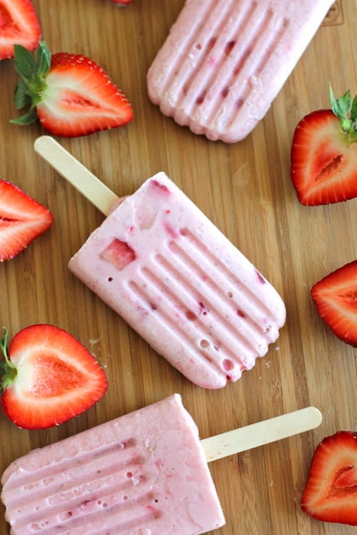 Strawberries and cream popsicles on a wooden surface, surrounded by fresh halved strawberries.