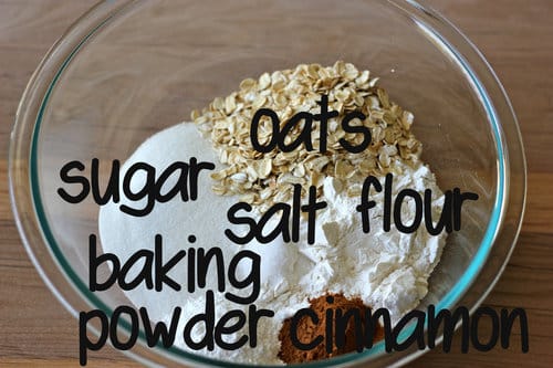 Glass mixing bowl filled with dry muffin ingredients including oats, flour, sugar, cinnamon, baking powder, and salt, labeled for baking preparation.