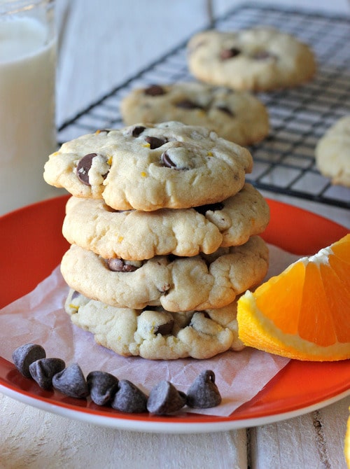 Stack of orange chocolate chip cookies on a red plate with chocolate chips and a fresh orange wedge, with more cookies and a glass of milk in the background.