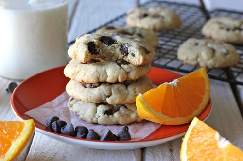Stack of orange chocolate chip cookies on a red plate with orange wedges and chocolate chips, with a cooling rack and glass of milk in the background.