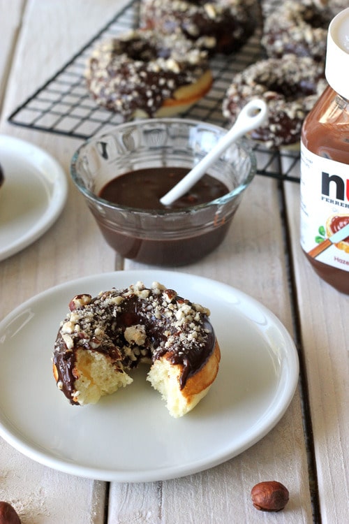 Nutella donuts topped with crushed hazelnuts, one donut with a bite taken out, next to a bowl of chocolate glaze.