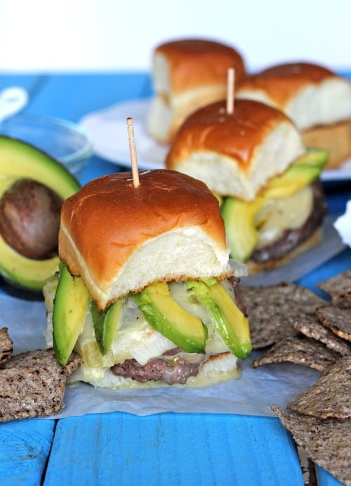 Stuffed green chili con queso cheeseburger sliders topped with avocado slices on soft slider buns, served with multigrain chips.