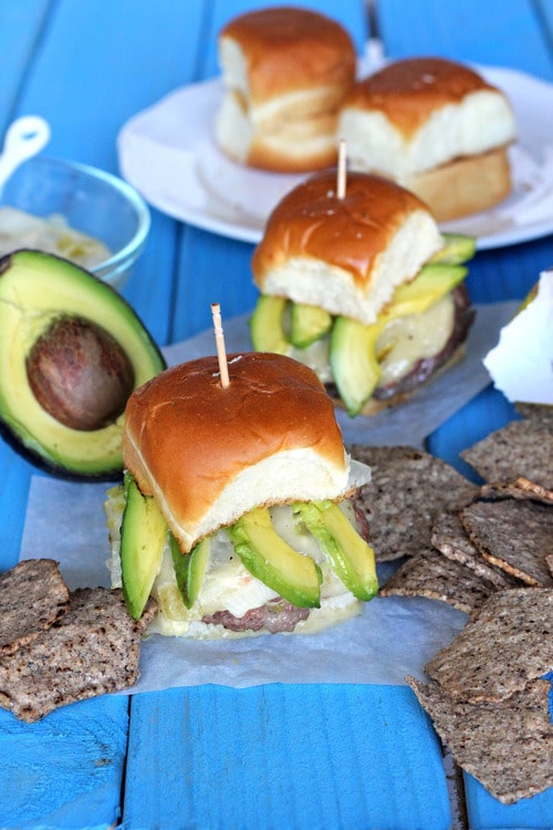 Cheeseburger sliders topped with sliced avocado on soft brioche buns, secured with toothpicks and served on a blue wooden table with crackers and a halved avocado nearby.