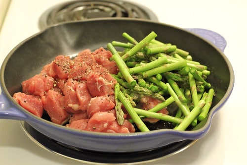 Raw diced pork and chopped asparagus seasoned with pepper in a skillet on a stovetop, prepared for cooking.
