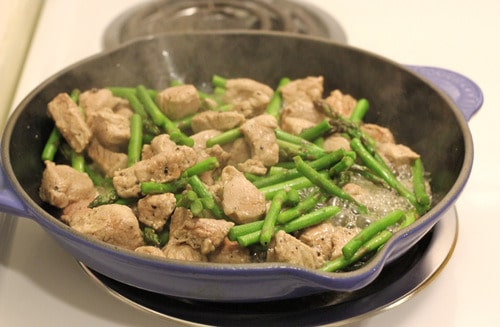 Pork and asparagus cooking in a skillet on the stovetop, with steam rising from the dish.