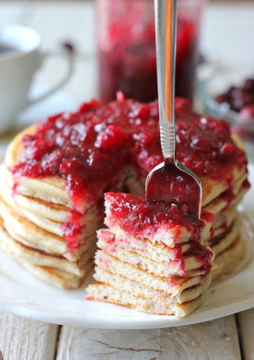 Stack of whole wheat pancakes topped with cranberry maple syrup, with a fork lifting a bite-sized portion from the stack.