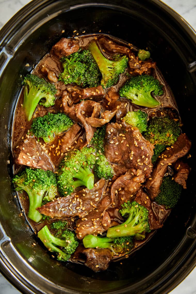 Slow cooker beef and broccoli in dark sauce while cooking, garnished with sesame seeds in a white bowl.