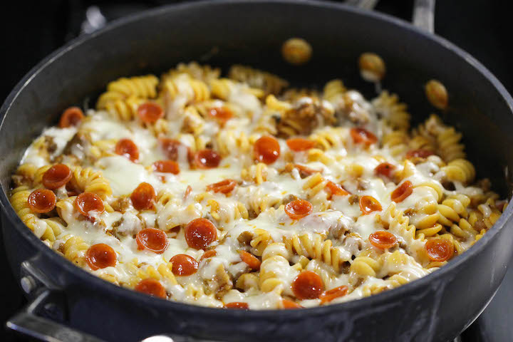One-pot pizza pasta bake with rotini, mini pepperoni slices, sausage, and melted mozzarella cheese in a black skillet.