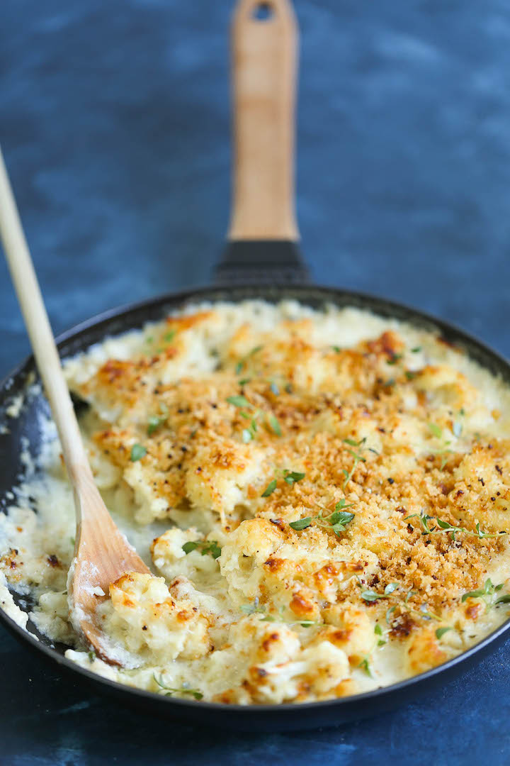 Creamy cauliflower gratin topped with golden breadcrumbs and fresh herbs in a black skillet with a wooden spoon.